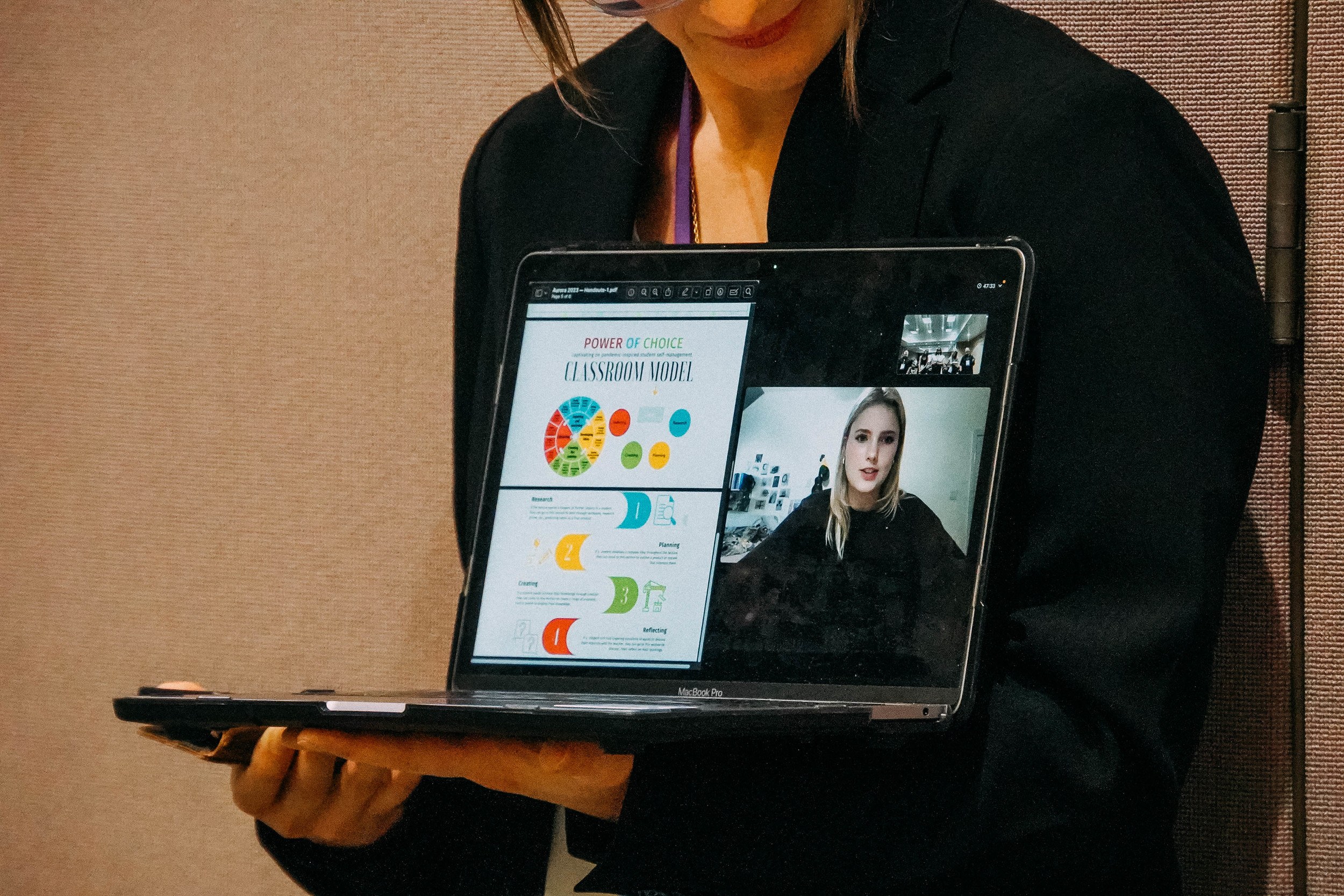 Presenter holding up a laptop, on which you can see a student talking and sharing a graphic.